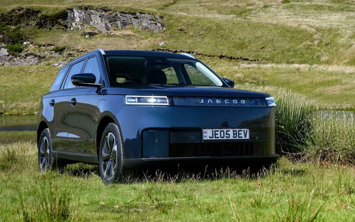 A dark blue Jaecoo J5 BEV parked on a grassy hillside, charge port on the front fender
